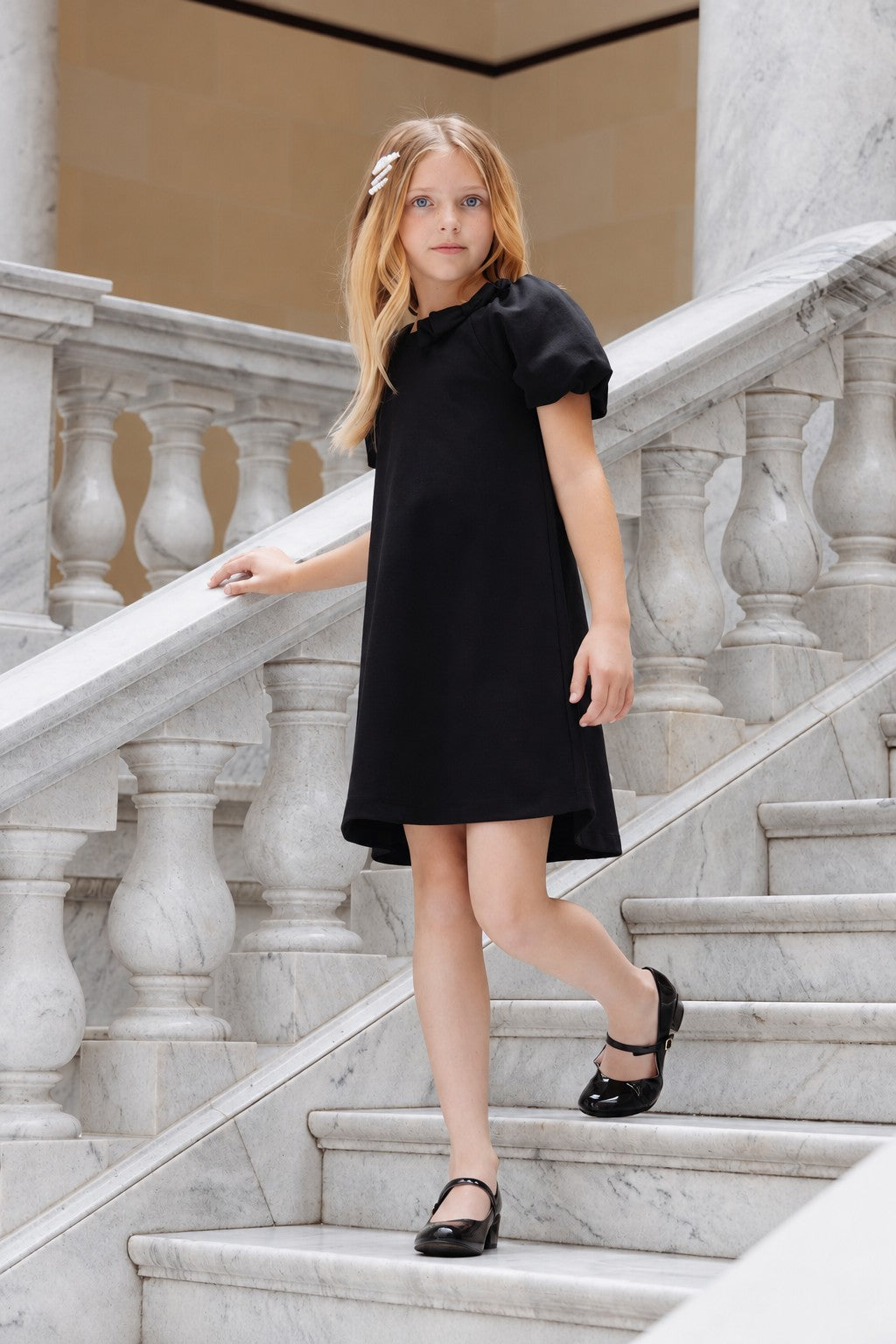 A young girl wearing a black bubble sleeve dress walks down a marble staircase. The dress features a relaxed silhouette and soft fabric, perfect for stylish, eco-friendly play.