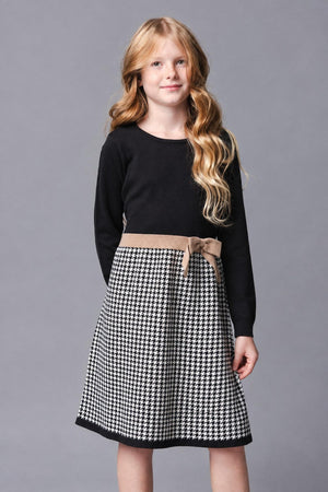 A young girl models a black long-sleeve dress featuring a houndstooth patterned skirt with a decorative beige bow at the waist. The dress is made from soft organic cotton.
