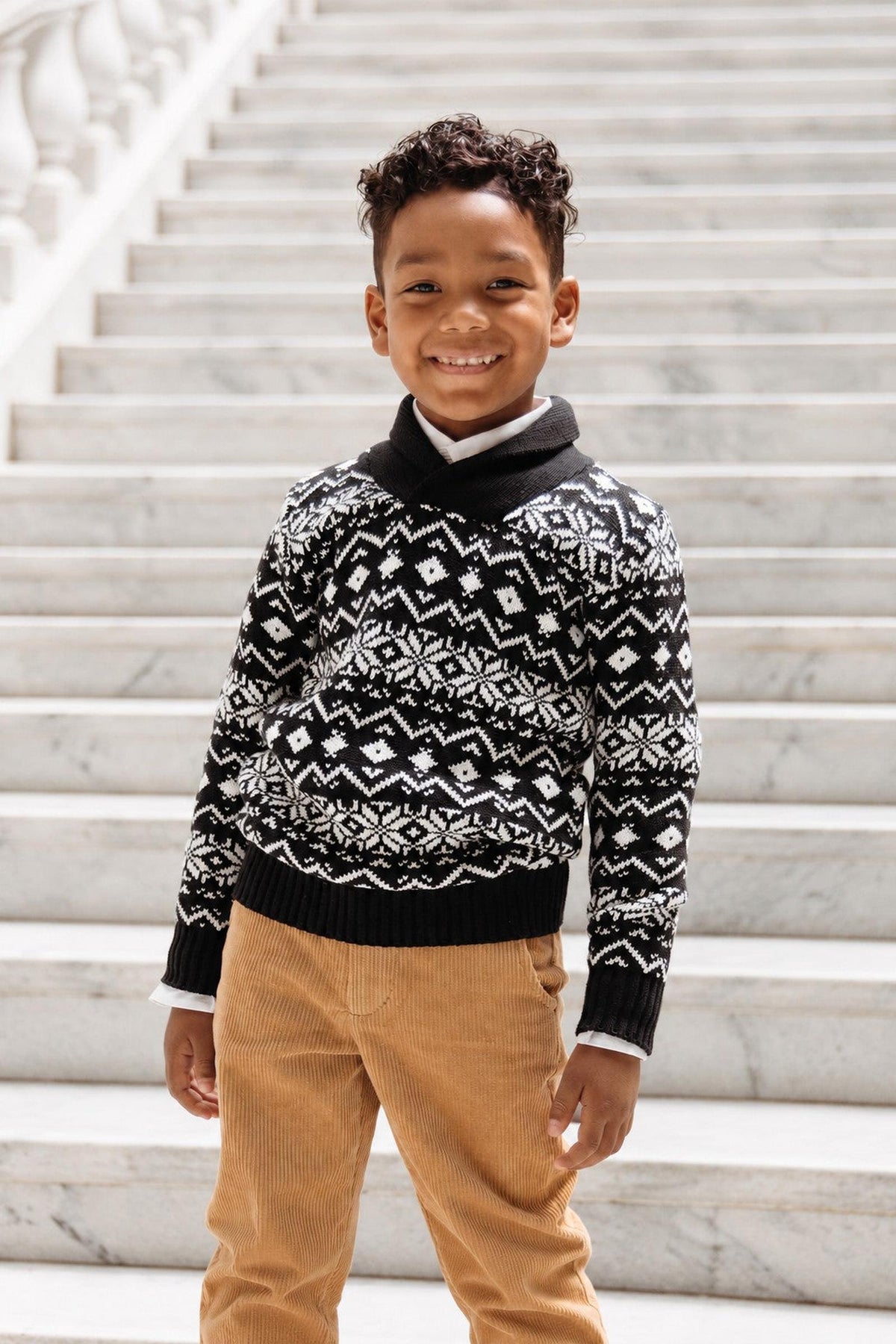 A young boy stands on marble steps, wearing a black and white patterned shawl collar sweater made from organic cotton. He smiles confidently, paired with beige corduroy pants.