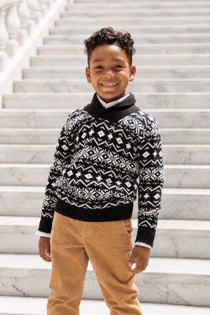 A young boy stands on marble steps, wearing a black and white patterned shawl collar sweater made from organic cotton. He smiles confidently, paired with beige corduroy pants.
