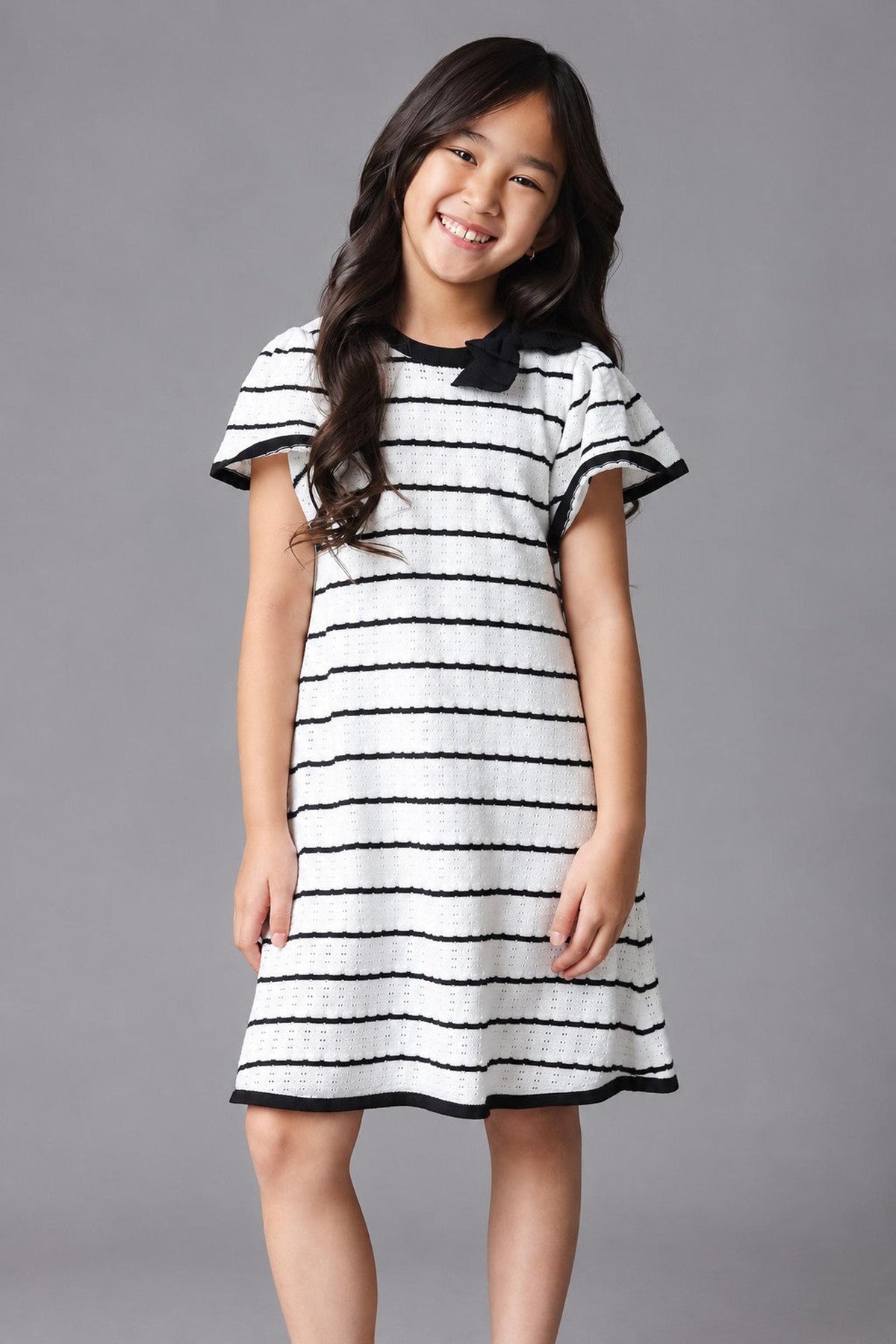 A young girl smiles while wearing a black and white striped organic cotton sweater dress with short sleeves and a bow detail at the neckline, set against a gray background.