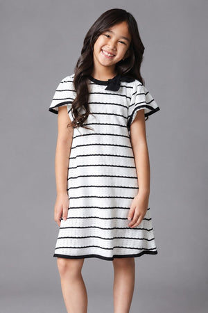 A young girl smiles while wearing a black and white striped organic cotton sweater dress with short sleeves and a bow detail at the neckline, set against a gray background.