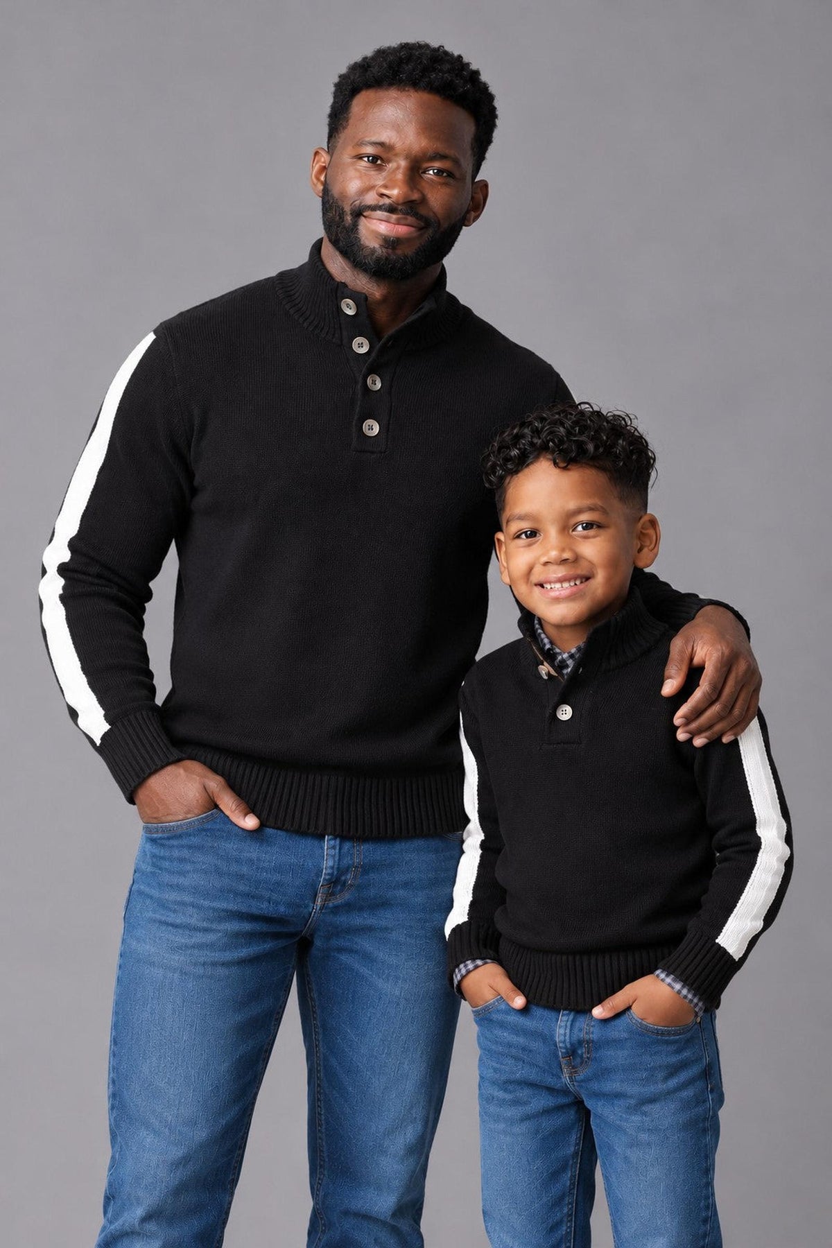 A father and son wear matching black mock neck sweaters featuring white stripes down the outer sleeves. The cozy, sustainable sweaters are styled with denim jeans against a gray backdrop.