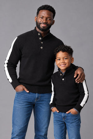 A father and son wear matching black mock neck sweaters featuring white stripes down the outer sleeves. The cozy, sustainable sweaters are styled with denim jeans against a gray backdrop.
