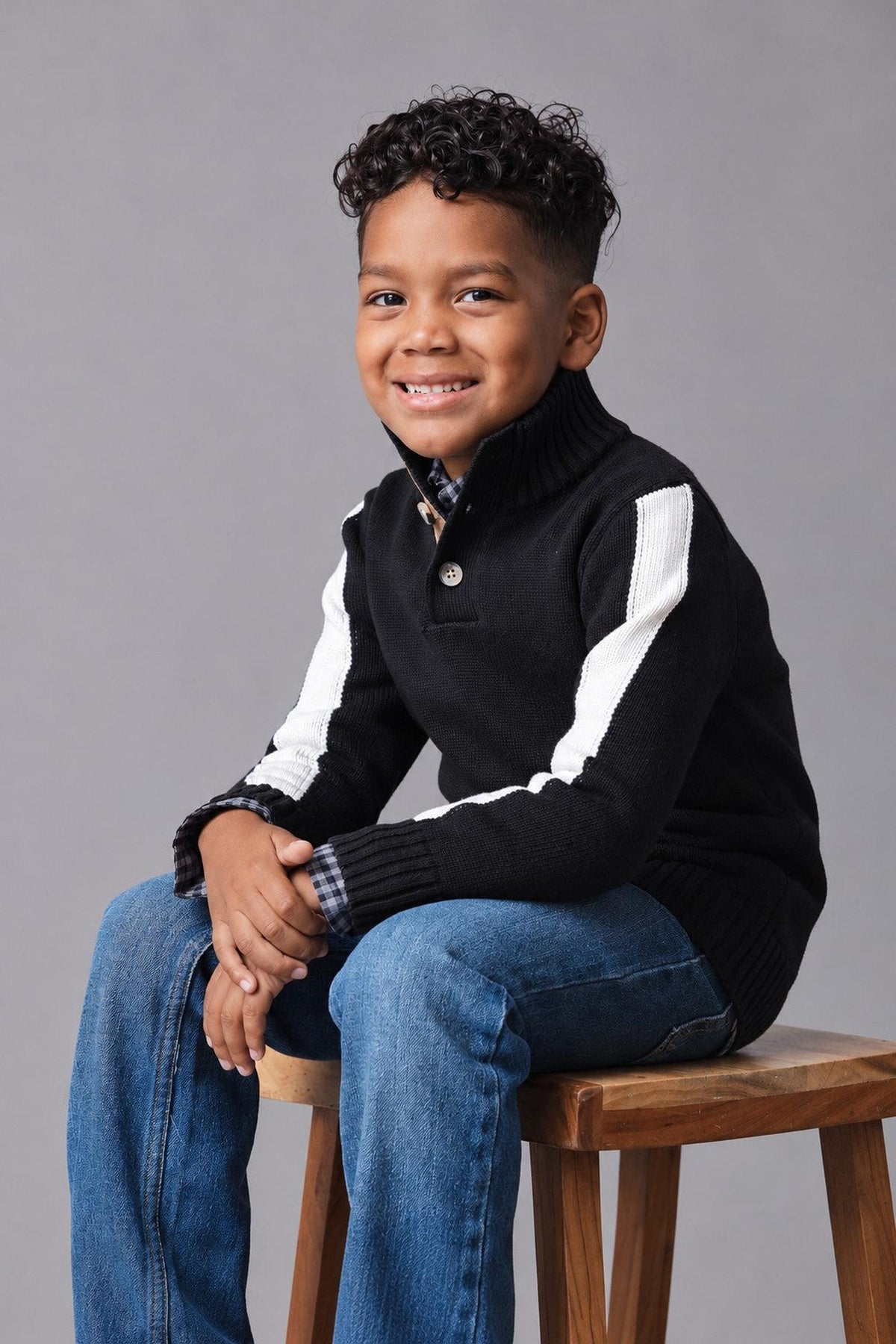 A young boy sits on a wooden stool, wearing a black mock neck sweater with white stripes on the sleeves and a faux suede button placket, paired with blue jeans.