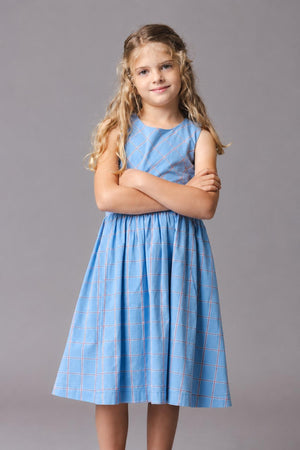 A young girl poses confidently in a sleeveless blue plaid dress, showcasing a fit-and-flare silhouette. The dress features a gathered waist and is made from organic cotton for comfort.
