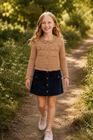 A smiling girl walks along a sunlit path, wearing a tan pointelle knit cardigan with ruffle trim and a navy skirt. She pairs the outfit with light pink sneakers.