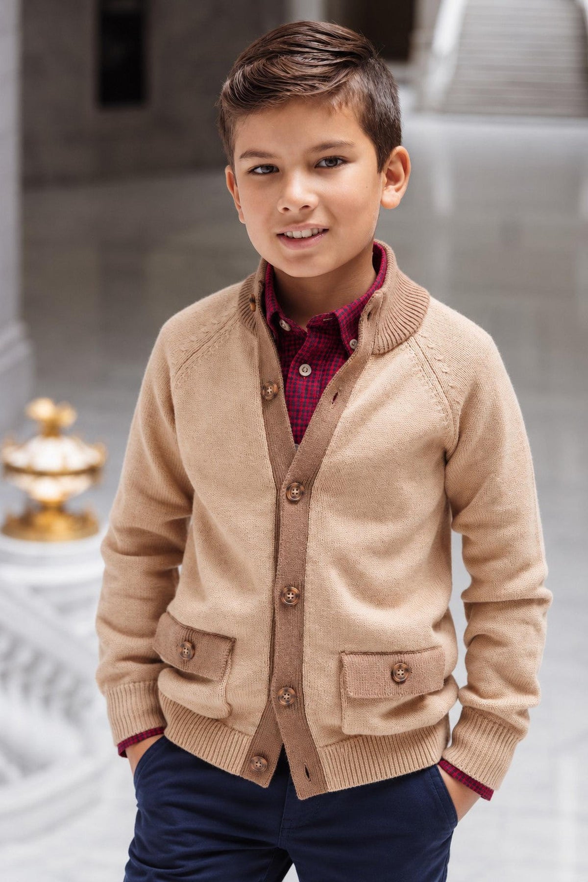 A young boy poses in a stylish camel-colored cardigan with a mock neck and front flap pockets, paired with a plaid shirt. The setting features elegant architecture in the background.