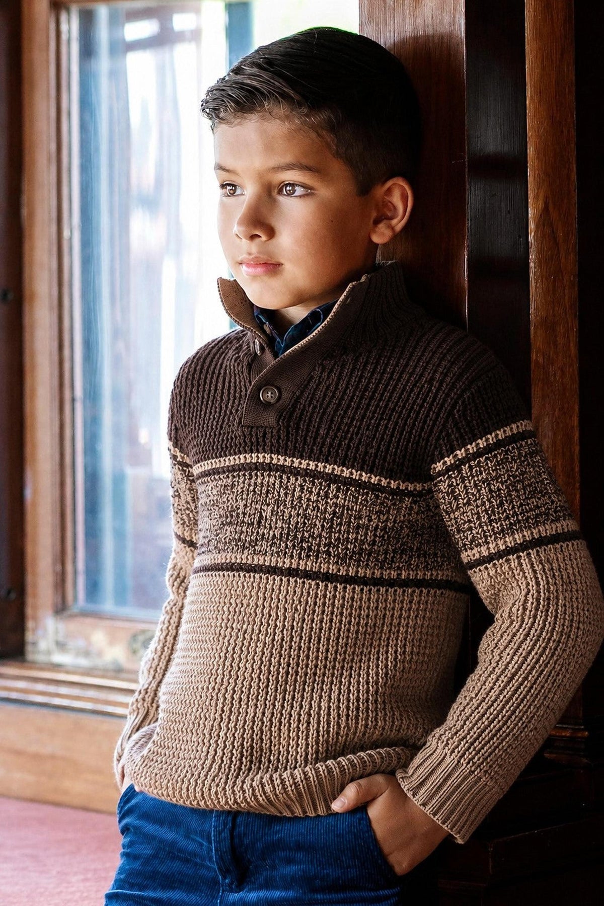 A boy stands by a window, wearing a stylish mock neck sweater featuring a textured waffle knit in camel and chocolate colors. The sweater is made from soft organic cotton.