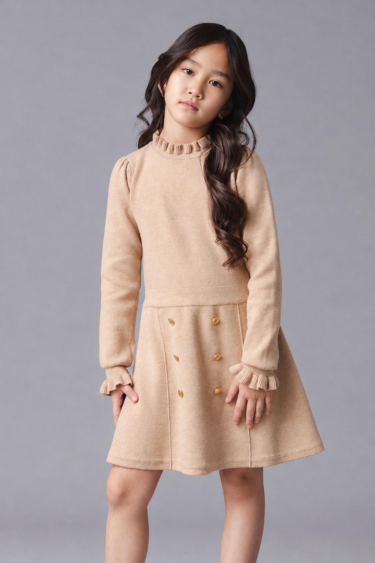 A young girl poses in a camel-colored organic cotton sweater dress featuring ruffle edges and gold buttons on the skirt.