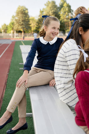 A young girl sits on a bleacher wearing a navy long-sleeve shirt with a white collar and camel-colored ponte pants.