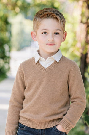 A young boy stands outdoors wearing a soft camel-colored V-neck sweater and a collared shirt. The setting features natural greenery, highlighting the cozy, timeless style of the outfit.