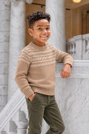 A smiling child stands by a marble staircase, wearing a camel-colored organic cotton sweater featuring a Fair Isle design.