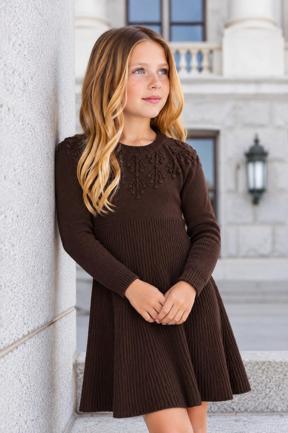 A young girl stands against a gray stone wall, wearing a chocolate heather sweater dress with a ribbed skirt and decorative shoulder stitching, showcasing a stylish and comfortable design.