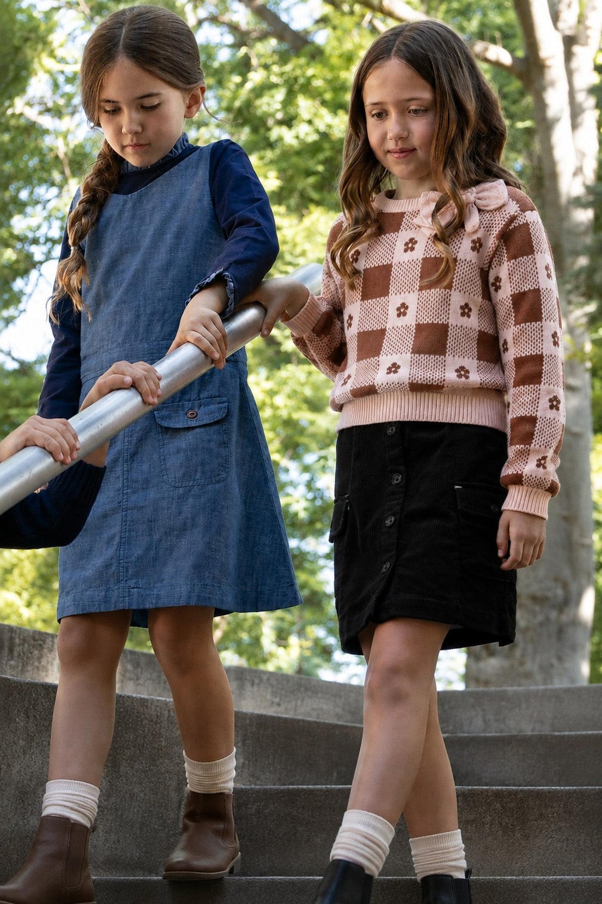 Two girls descend a staircase in a park. One wears a denim dress, while the other sports a pink and brown intarsia sweater, showcasing a playful design and cozy style.
