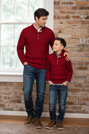 A father and son stand together in a cozy setting, both wearing matching red organic cotton mock neck cable sweaters.