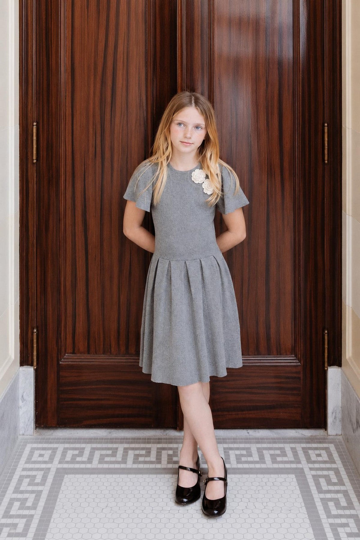 A young girl stands confidently in a dark gray organic cotton pleated sweater dress, featuring a floral rosette at the neckline.