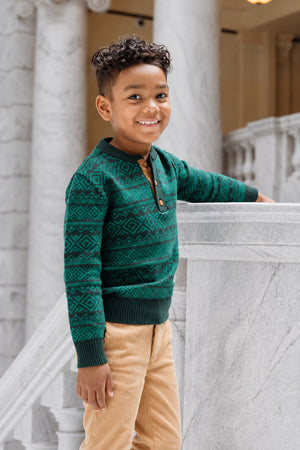 A young boy smiles while leaning against a marble railing, wearing a deep green organic cotton sweater with a patterned design and a henley collar.