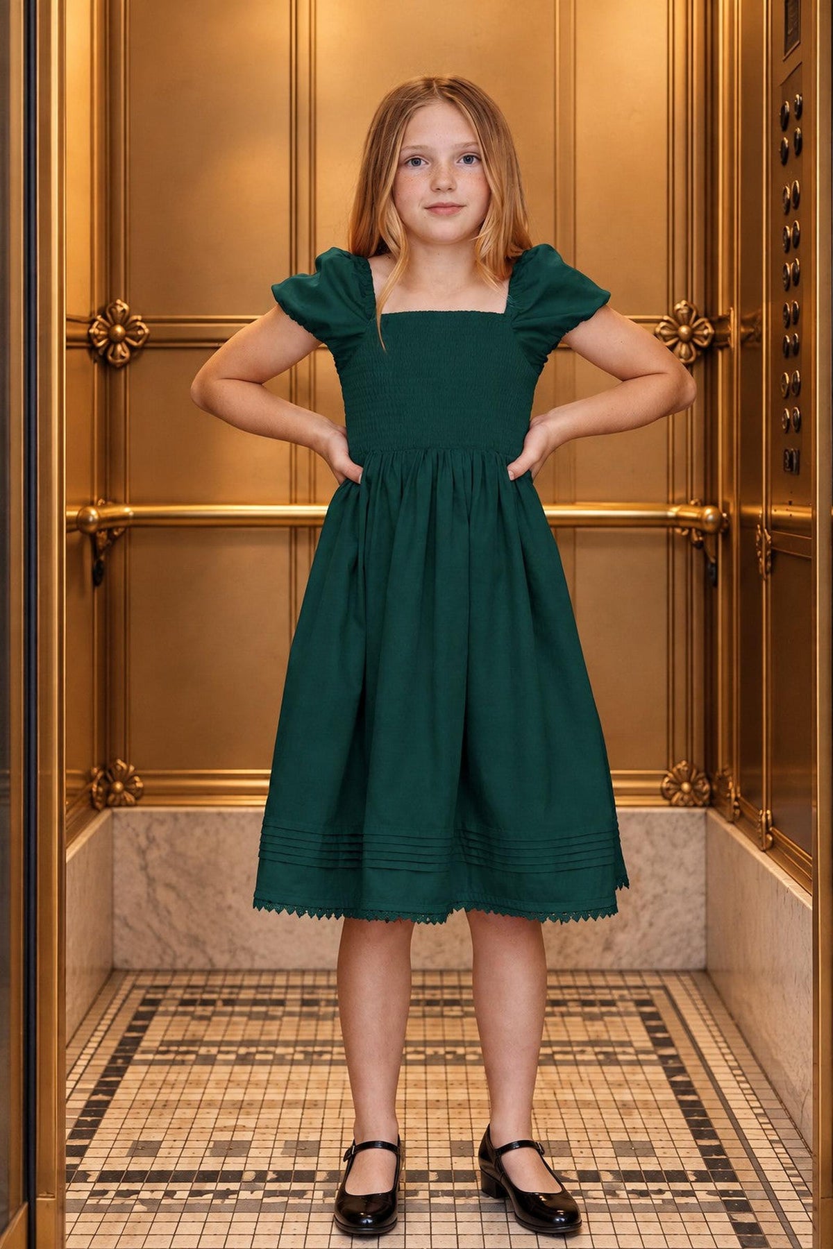 A young girl poses confidently in a deep green organic cotton sateen dress with a smocked bodice and puffed sleeves, set against a luxurious gold elevator backdrop.