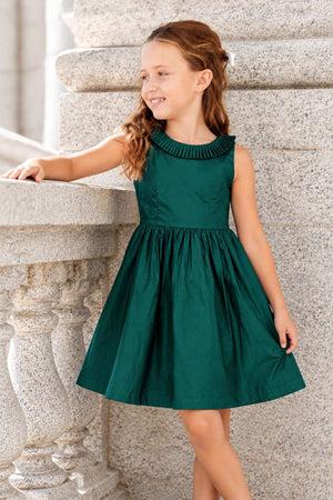 A young girl stands smiling beside a stone railing, wearing a deep green organic cotton party dress with a pleated collar and a full skirt, perfect for festive occasions.