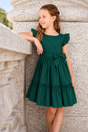 A girl stands beside a stone railing, wearing a deep green organic cotton tiered party dress with flutter sleeves and a waist sash bow, showcasing a playful and festive style.