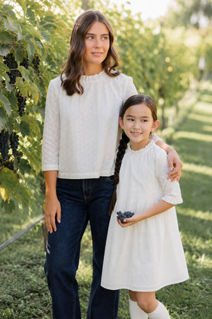 A woman and a girl stand together in a vineyard, both wearing ivory eyelet outfits. The woman wears a ruffle neck top, while the girl holds a bunch of grapes.