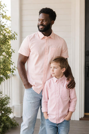 A man and a boy stand together outside, both wearing matching rose-colored seersucker shirts. The man has a beard and the boy looks up at him, both smiling.