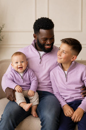 A man, a boy, and a baby sit together on a beige couch, all wearing matching lavender organic cotton half-zip sweaters. They smile warmly in a cozy, well-lit setting.