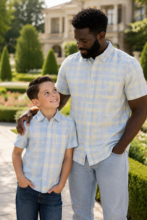 A man and a boy, both wearing light blue and yellow plaid short sleeve button-down shirts, smile at each other in a lush garden setting, showcasing their matching outfits.