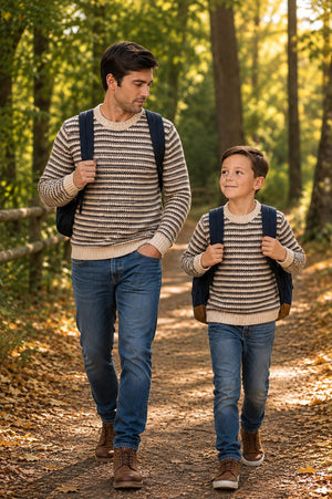 A man and a boy walk together on a forest path, both wearing matching brown and tan striped organic cotton crew neck sweaters. The setting features vibrant autumn foliage.