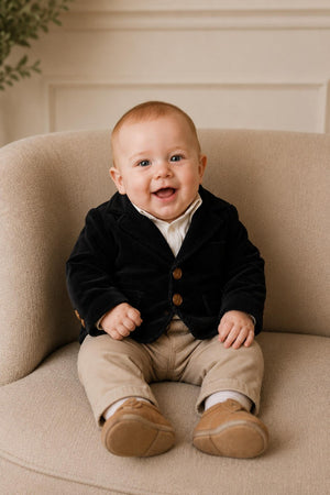A smiling baby sits on a neutral-colored sofa, wearing a black organic cotton corduroy blazer with wooden buttons and beige pants, showcasing a stylish and comfortable outfit for toddlers.