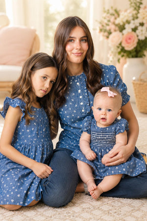 A woman sits on a rug with two young girls, all wearing matching blue chambray outfits with puff sleeves. The setting features soft lighting and floral decor in the background.