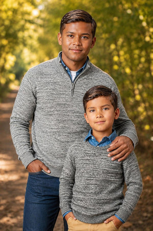 A man and a boy stand outdoors in a wooded area, both wearing matching gray organic cotton half-zip pullovers. The setting features dappled sunlight filtering through the trees.