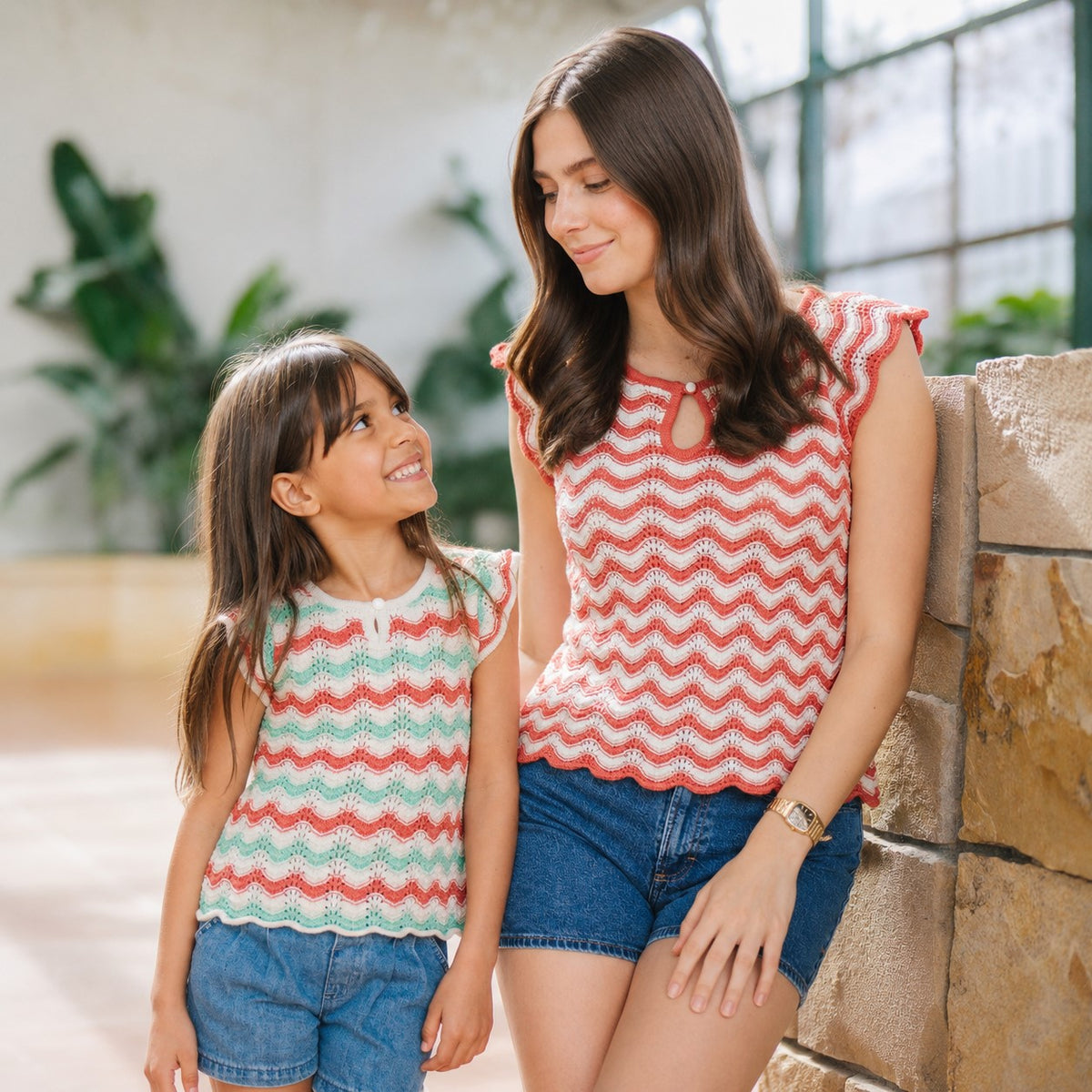 A woman and a girl are smiling at each other, wearing matching organic cotton, wave-patterned sweater tops in light green and red. They are in a bright, tropical setting.
