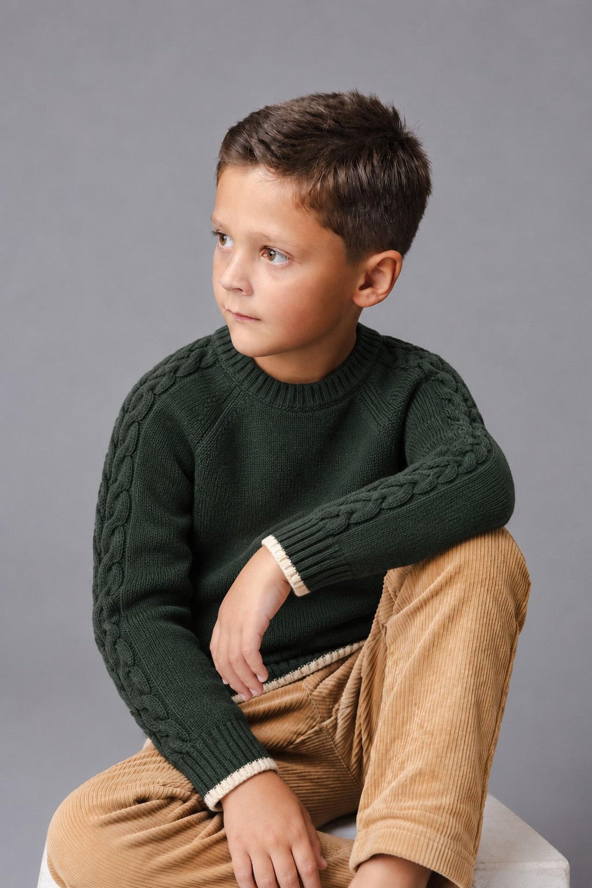 A young boy sits thoughtfully, wearing a forest green organic cotton sweater with cable detailing on the sleeves.