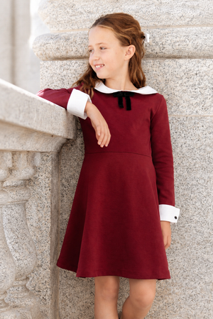 A young girl stands against a stone pillar, wearing a stylish oxblood dress with a white Peter Pan collar and bow. The dress features long sleeves and a flared skirt.