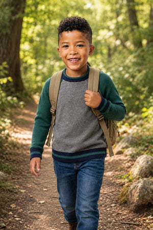 A young boy is walking along a forest path, wearing a gray and green organic cotton raglan sweater. He carries a beige backpack and smiles happily amidst the greenery.