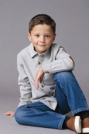 A young boy sits casually, wearing a grey organic cotton flannel shirt with button details and elbow patches. He pairs it with blue jeans, showcasing a relaxed and stylish look.