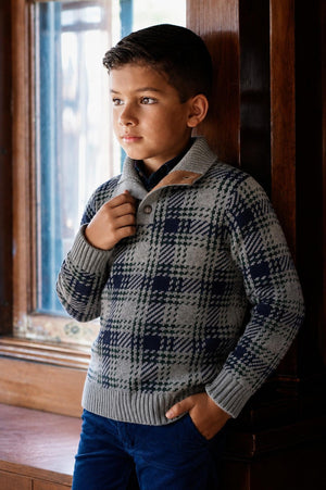 A boy stands by a window, wearing a gray and navy plaid mock neck sweater with a button detail.