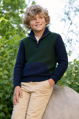 A smiling boy with curly hair is wearing a navy and green half-zip sweater made from organic cotton, paired with light beige pants, while standing on a large rock in.