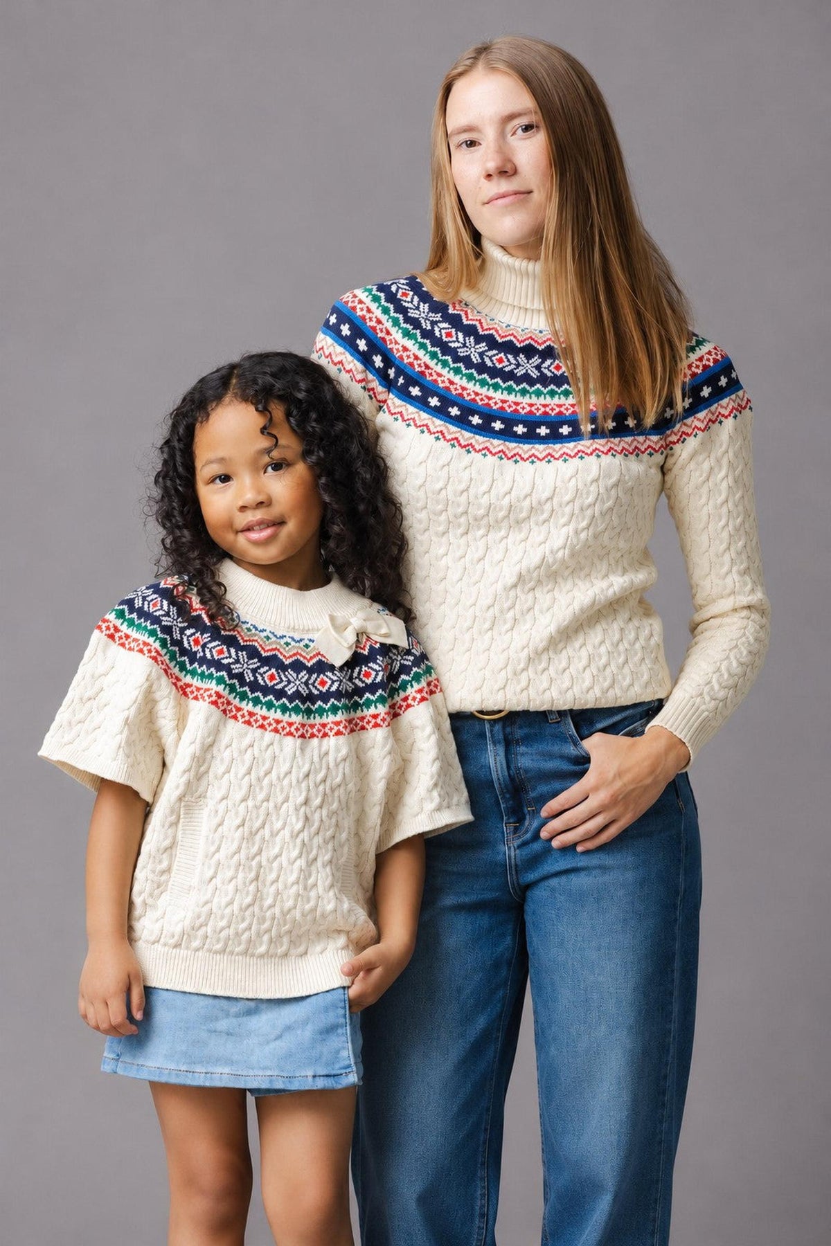 A woman and a girl stand side by side, both wearing matching ivory cable sweaters with a colorful fair isle pattern at the shoulders.