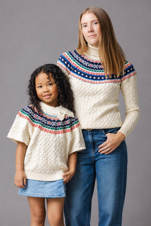 A woman and a girl stand side by side, both wearing matching ivory cable sweaters with a colorful fair isle pattern at the shoulders.