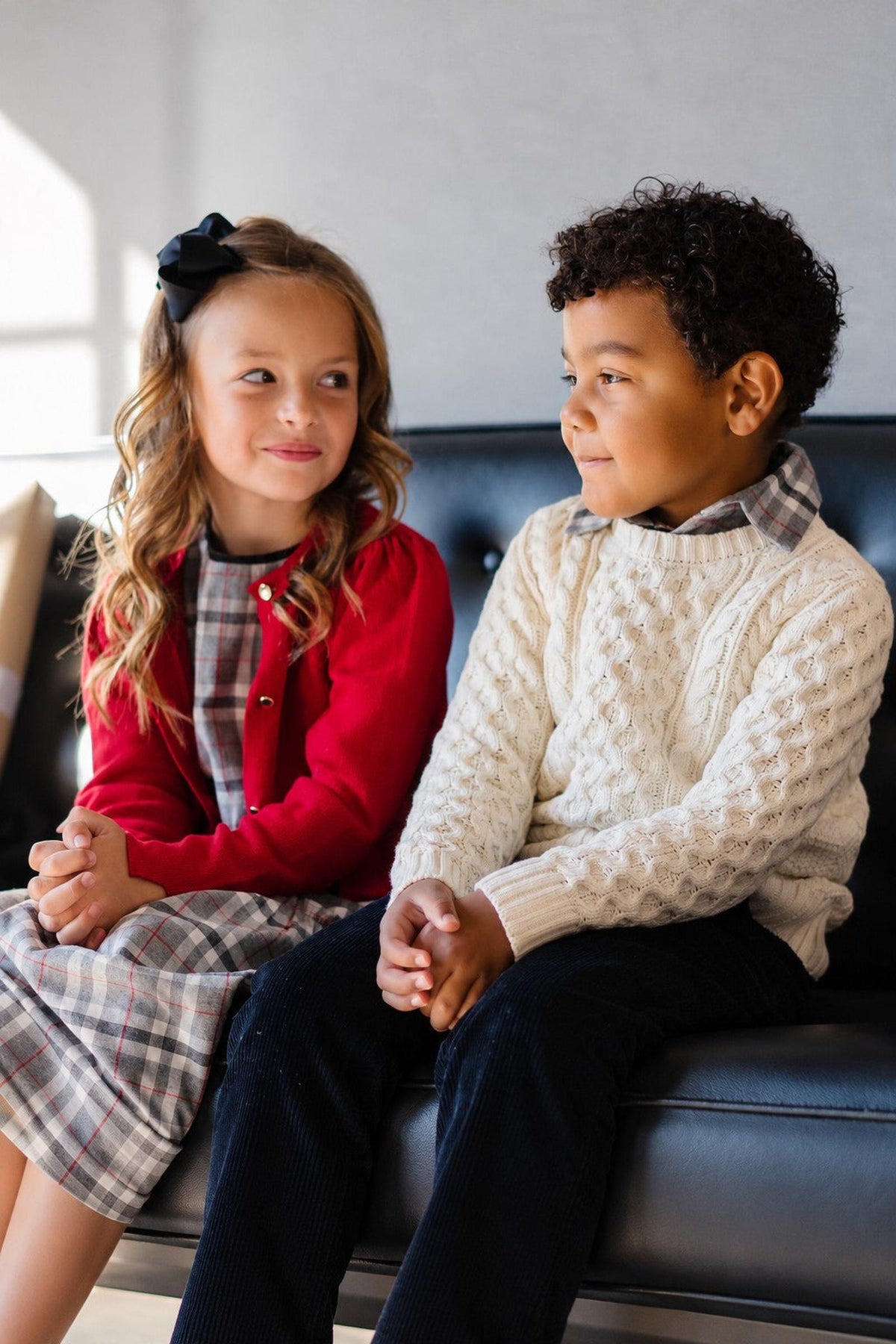 A girl in a red cardigan and a boy in an ivory cable knit sweater sit together on a black couch, smiling at each other in a bright, modern setting.