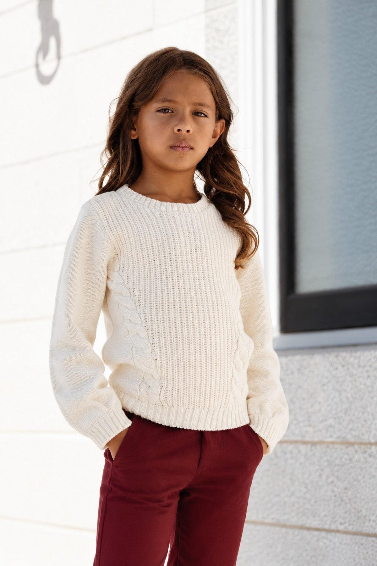 A young girl poses confidently in an ivory cable knit sweater with bubble sleeves, paired with burgundy pants. The sweater showcases a cozy texture, perfect for casual, stylish outfits.