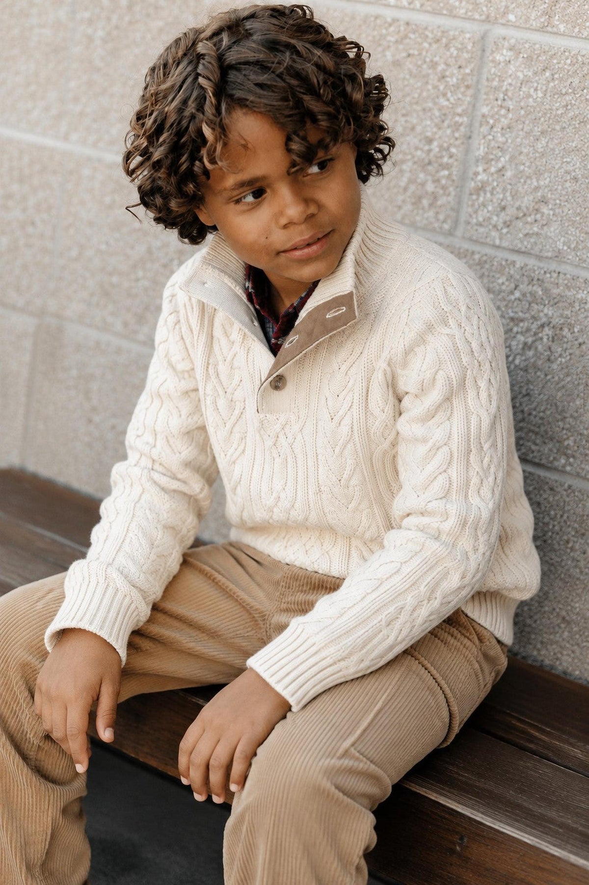 A young boy with curly hair sits on a wooden bench, wearing a cream-colored cable knit sweater with a mock neck and button detail.