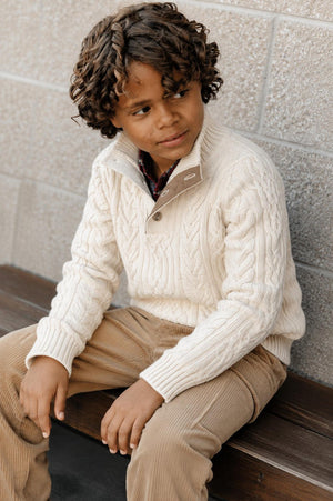 A young boy with curly hair sits on a wooden bench, wearing a cream-colored cable knit sweater with a mock neck and button detail.