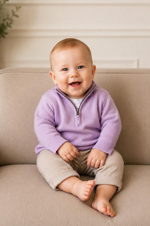 A smiling baby sits on a beige sofa, wearing a lavender organic cotton half zip sweater and light beige pants.