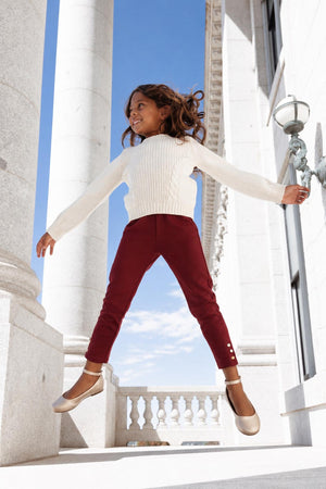 A young girl jumps joyfully in a bright setting, wearing a cozy cream sweater and oxblood ponte pants with gold button cuffs. She complements the outfit with beige ballet flats.