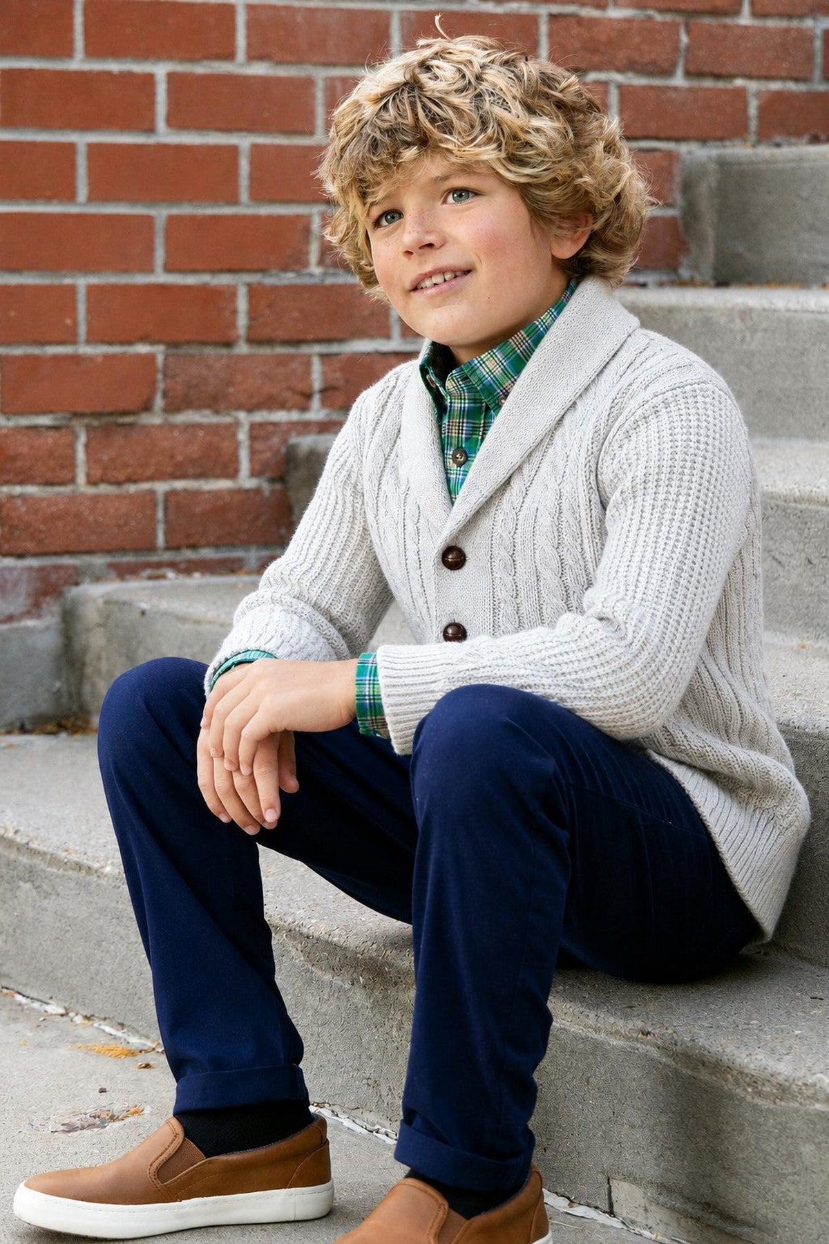 A young boy sits on stone steps, wearing a light gray shawl collar cardigan made from organic cotton, paired with a plaid shirt and navy pants, showcasing a classic and.