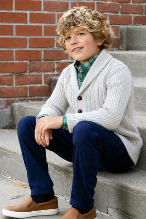 A young boy sits on stone steps, wearing a light gray shawl collar cardigan made from organic cotton, paired with a plaid shirt and navy pants, showcasing a classic and.